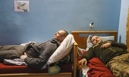 Two soldiers in uniform lying on top of beds perpendicular to each other, one with his hand on his chest and looking at the camera, with a child’s drawing of a bird on the wall