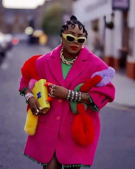 Portrait of Sade Adeyemi wearing a pink overcoat with green top, gold necklace, yellow sunglasses, yellow handbag and some red, green and purple fluffy things under her arm.