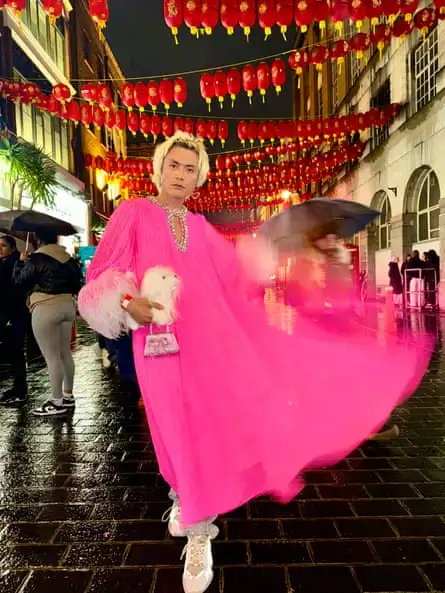 Yu Masui in Chinatown, London, wearing a bright pink long dress, with diamante trim at the neck and white fluffy cuffs. He is holding a fluffy toy animal and sparkling mini handbag.