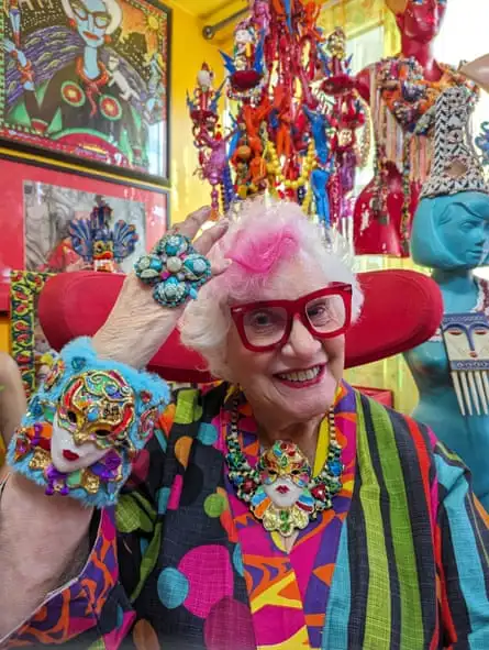 Sue Kreitzman. There’s a lot of colours and stripes. She’s wearing red-framed glasses and on her wrist is a large wrap with a small theatrical mask attached.