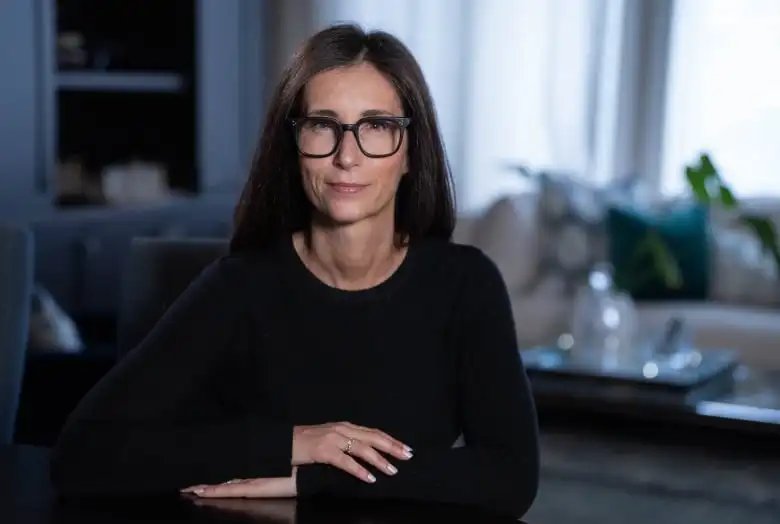 A woman with long hair, seated wearing a black top and black glasses.