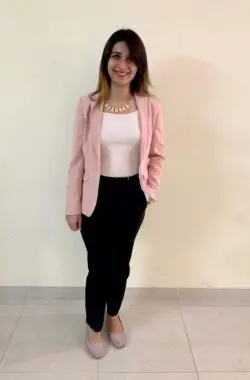 A woman poses against a white wall wearing a pink blazer and black pants.