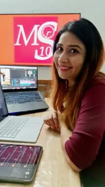 A woman sits at a desk with two laptops and a tablet open in front of her. A poster is visible in the background and says