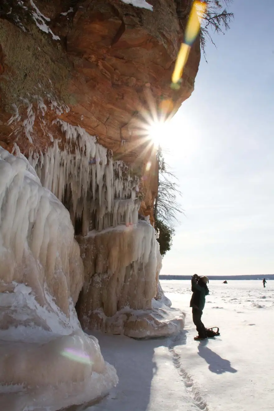 the sun shines behind a cliff covered in frozen ice. figures stand below and look up in wonder