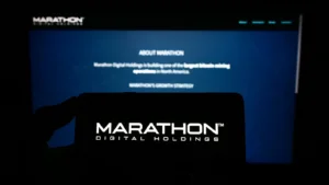 Person holding mobile phone with logo of American company Marathon Digital Holdings Inc. on screen in front of web page. Focus on phone display. Unmodified photo. MARA stock