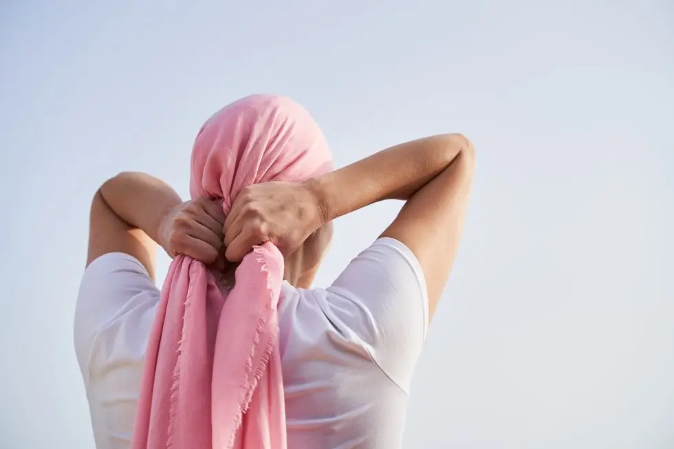 unrecognizable female cancer patient putting a pink scarf on her head with the sky in the background