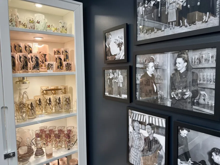 A cupboard of bright pink glassware sits next to a wall of black and white photos. The photos show the women employees of Gay Fad Studios through the years.