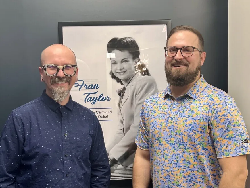 Two men stand smiling in front of a black and white poster of Fran Taylor, a glassware pioneer of the 1940s.