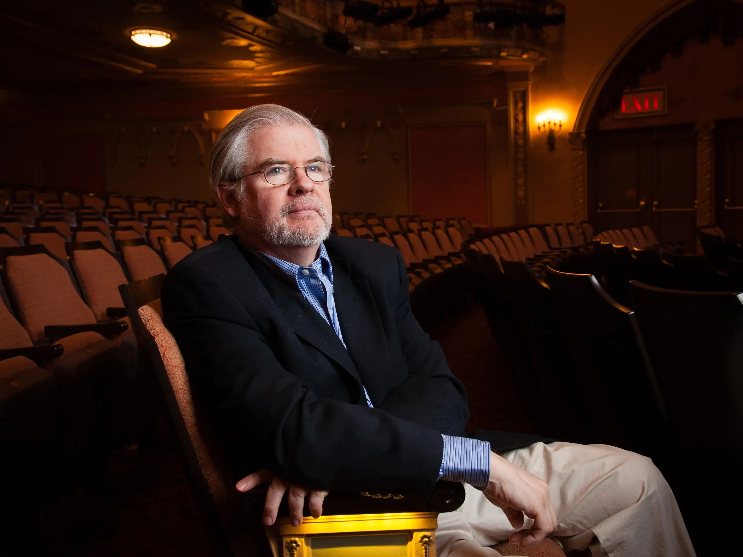 Late playwright Christopher Durang sitting in a theatre auditorium. Durang wears khaki pants a blue buttonup shirt and a…