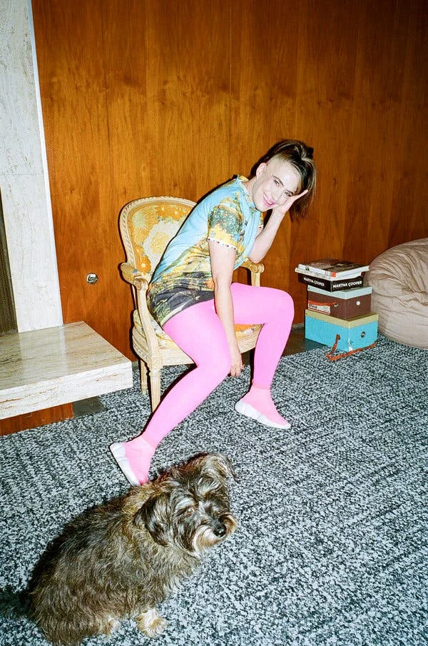 A woman in pink leggings and a light blue shirt with a graphic printed on it sits in a gold chair and rests her head on her hand. A pile of books, a rug and a dog are positioned around her.