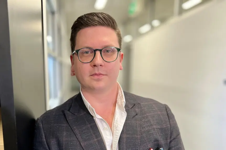 A man in a blazer and glasses looks at the camera.