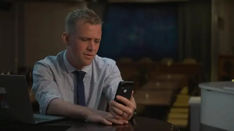 A man in a dress shirt and tie looks down at a cell phone.