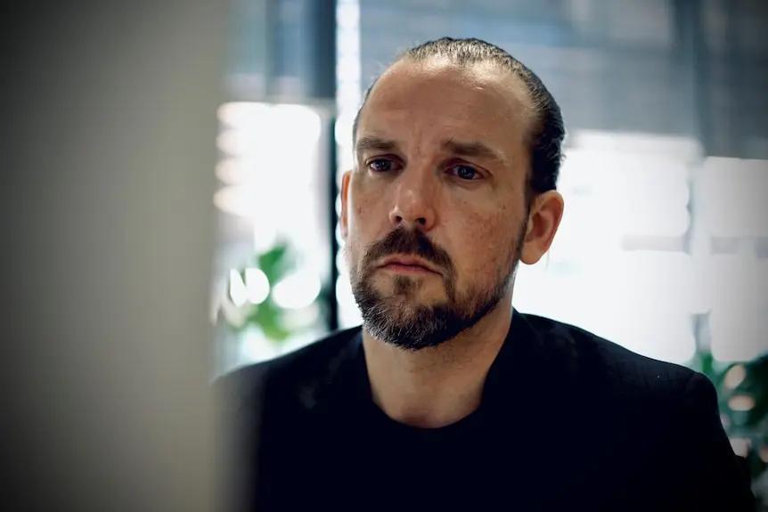 A man with a dark trimmed beard and his hair pulled back into a bun looks at a computer screen in an office.