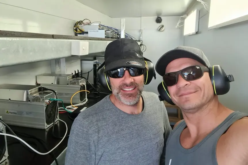 Two men wearing industrial ear muffs, black protective glasses and caps in a small room with electrical equipment.