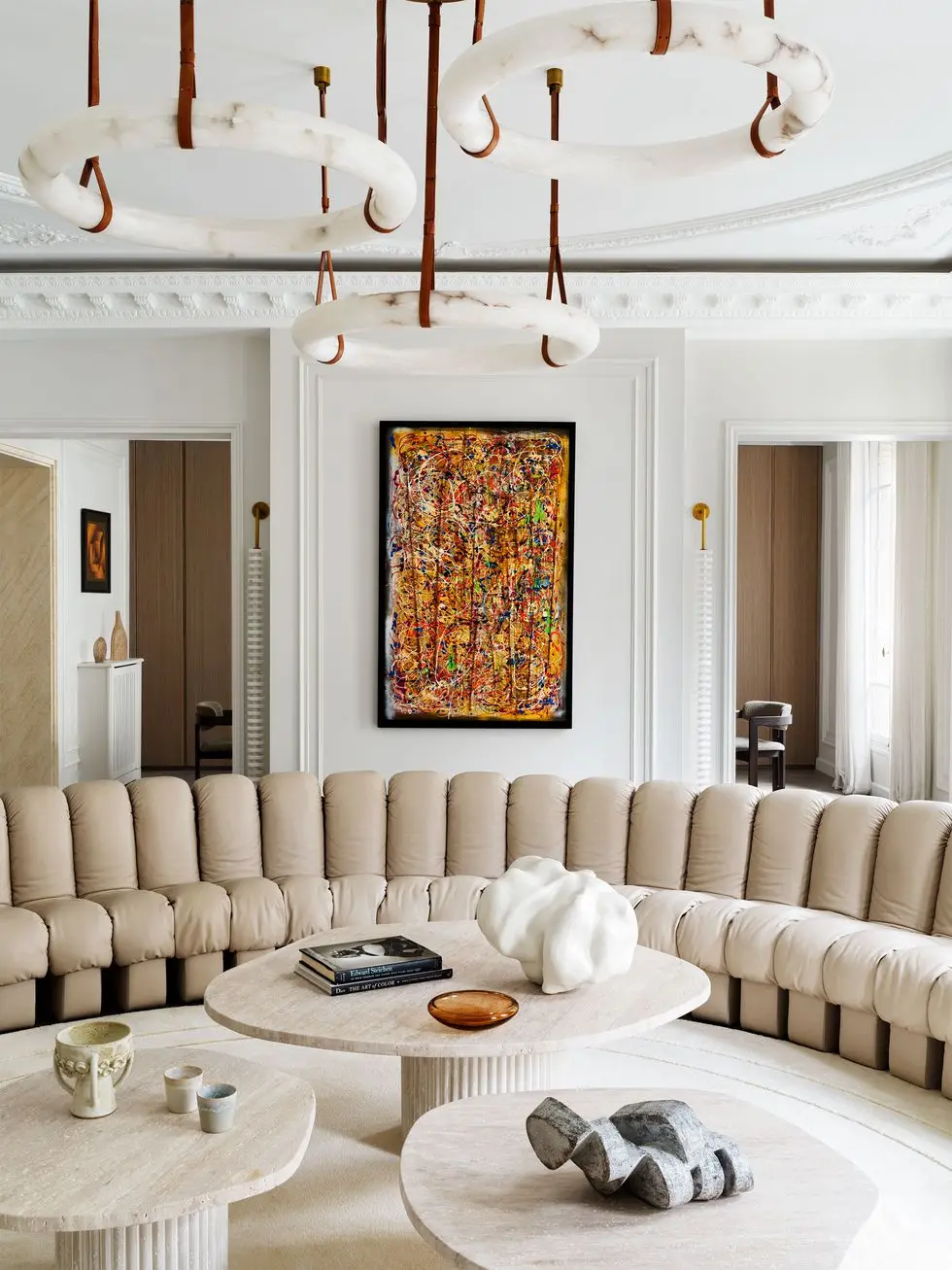 semicircular sofa in beige tufted leather, three cocktail tables with sculptures, colorful splatter painting on wall, two doors leading to dining room, three circular alabaster pendants hung with leather straps