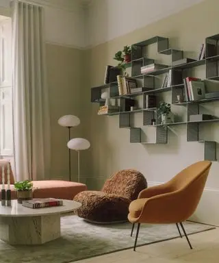 A living room with light green walls, sculptural hanging bookshelves and warm-toned furniture