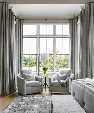 drapes vs blinds, gray bedroom with large bay window, grey floor to ceiling drapes, pair of armchairs in window, rug, bed to the right