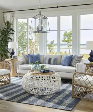 A white living room with large windows, a white couch with blue throw pillows, a round rattan coffee table with a tray with blue flowers on it, and a striped blue and white rug underneath it