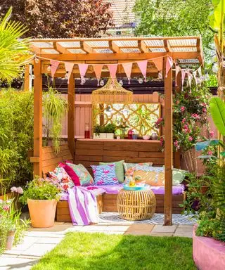 A rich wooden pergola with pink, blue, and yellow patterned throws and throw pillows underneath it, with a lawn in front of it and green trees around it