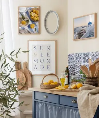 A kitchen with a white wall with three prints on the wall, a chopping area with blue and white mosaic tiles and a wooden sideboard with lemons on top of it and cornflower blue drawers and curtains underneath