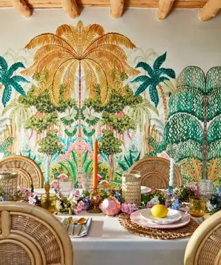 A dining room with tropical gold and green tree printed wallpaper, a rectangular white dining table with flowers, glasses, and plates on top, with four light rattan chairs on the sides of it