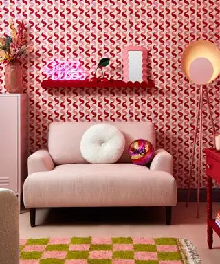 A living room with pink wavy wallpaper, a red scalloped shelf, a light pink armchair with a round throw pillow and disco ball on it, and a pink and green checked rug in front of it