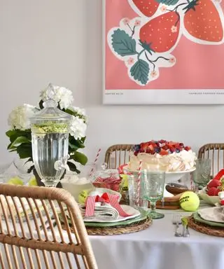 A dining room with a white tablecloth with a pavlova, plates, tennis balls, white flowers, and a glass trophy on top with three wooden rattan chairs around it and a pink strawberry print above it