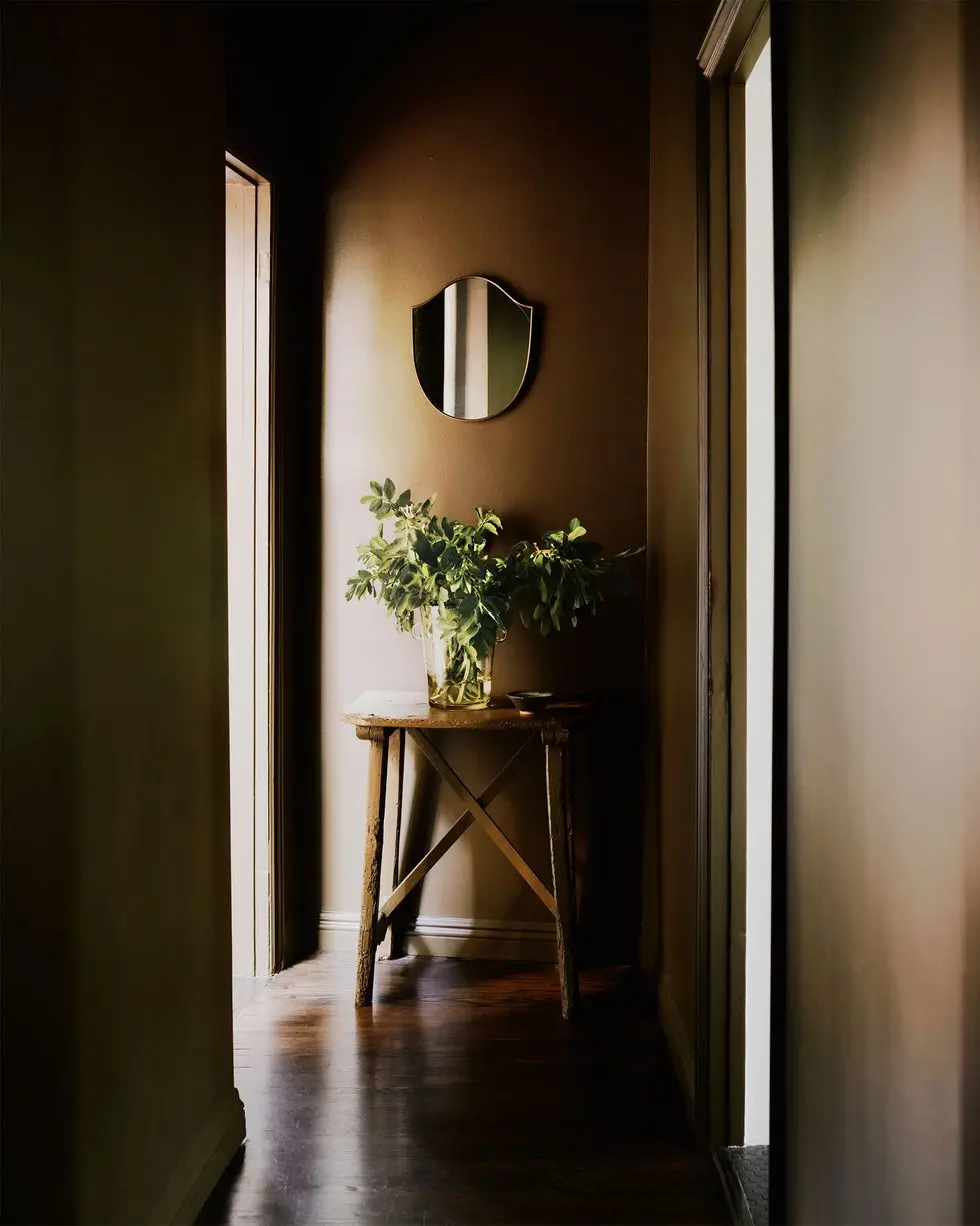 a dark entry all with wood floor, rustic wood stool with a glass vase and greenery, and a shield shaped mirror hanging above