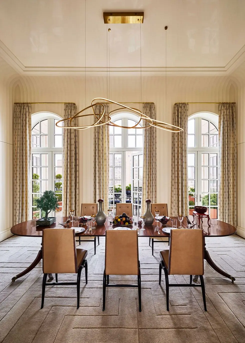 classic dining room with a oval table and chairs with three floor to ceiling windows at back with beige patterned to the floor draperies and a subtle monochrome dark beige carpet with a deconstructed greek key design