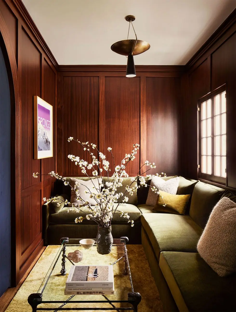 a wood paneled living room corner with a sofa in light green and a glass top cocktail table with a vase holding pretty branches