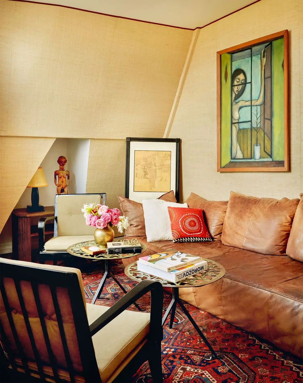 a small living room with a vintage leather sofa, two round metal cocktail tables, two wooden armchairs with cushions, painting of a woman, a nook with a small bookcase, table lamp, and wood statue, turkish rug