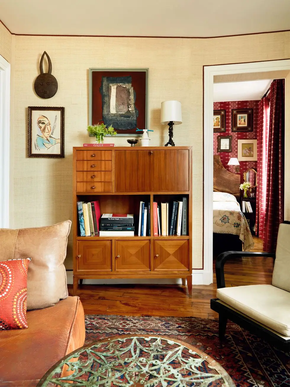 a leather sofa, cocktail table, chair with cushions, a wood storage cabinet with three small doors below, a shelf with books, and four small drawers and a pull down compartment at top, a lamp on top and artworks on wall