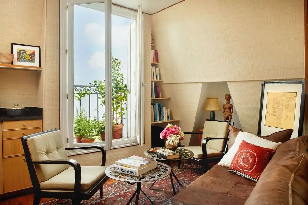 a vintage light brown leather sofa, two armchairs, two round bronze cocktail tables, a large window, with built in shelves on right and a chest of drawers on left opens to a small terrace with plants, nook with lamp and statue