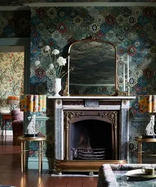 A living room with dark blue textured wallpaper, a fireplace with a gold arched mirror and white vase of flowers on top, two gold side tables with leopard lamps with yellow floral lampshades, and a coffee table