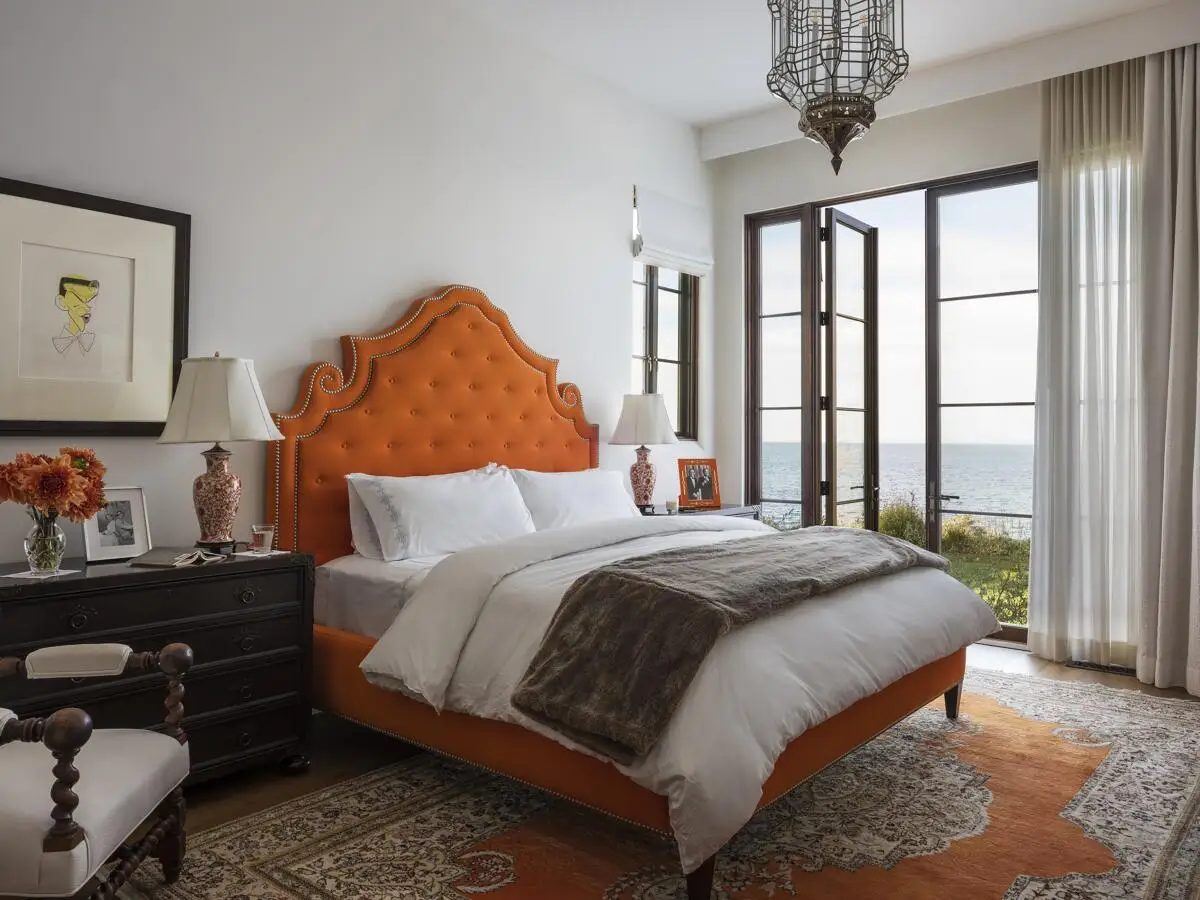 A bedroom has a curvaceous headboard upholstered in moleskin in orange, flanked by ginger-jar lamps.