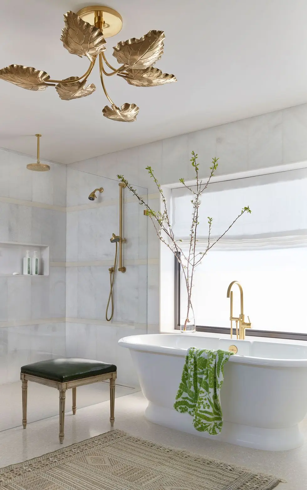 white bathroom with a stand alone tub with a gold tone fixtures and a green patterned towel draped over it and a gold leaf chandelier above