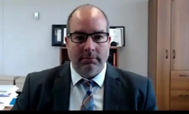 A bald man in a suit and tie is sitting in an office facing a camera, unsmiling.