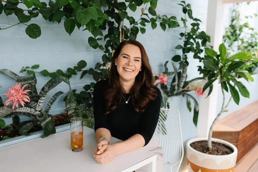 a woman with brown hair smiles sitting at a cafe table with plants surrounding her