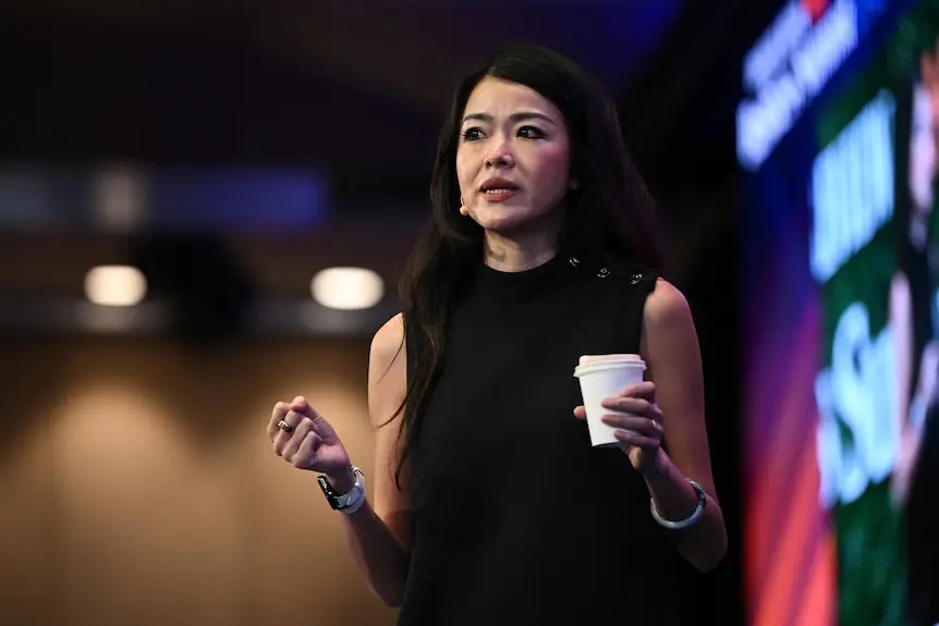 A mic'd-up middle-aged woman with long dark hair speaks while holding a coffee cup onstage at a meeting.