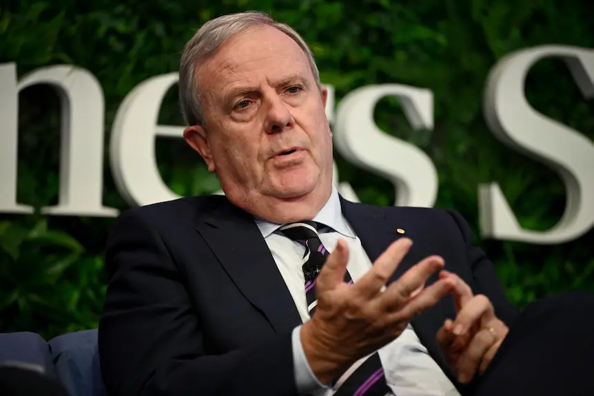 A man in a suit with grey hair leans back and gestures as he speaks on a stage.