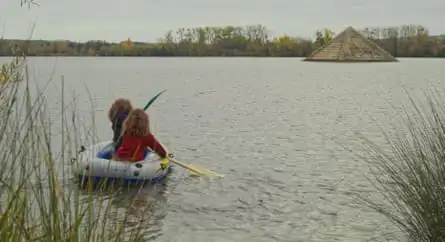 Two little girls paddle a dinghy across a lake towards a ziggurat shaped island