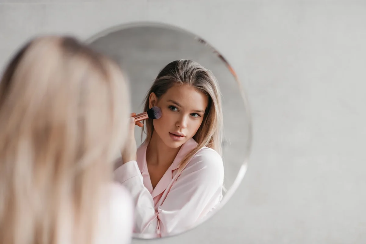 Beautiful woman in pajamas in front of mirror, holding brush and applying makeup, face powder or bronzer in bathroom, free space. Cosmetics for makeup concept