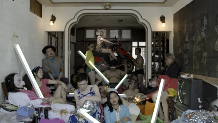 A film still showing a group of people, some dressed, some half naked, sit on the floor of a living room or pose in the doorway holding LED bars.