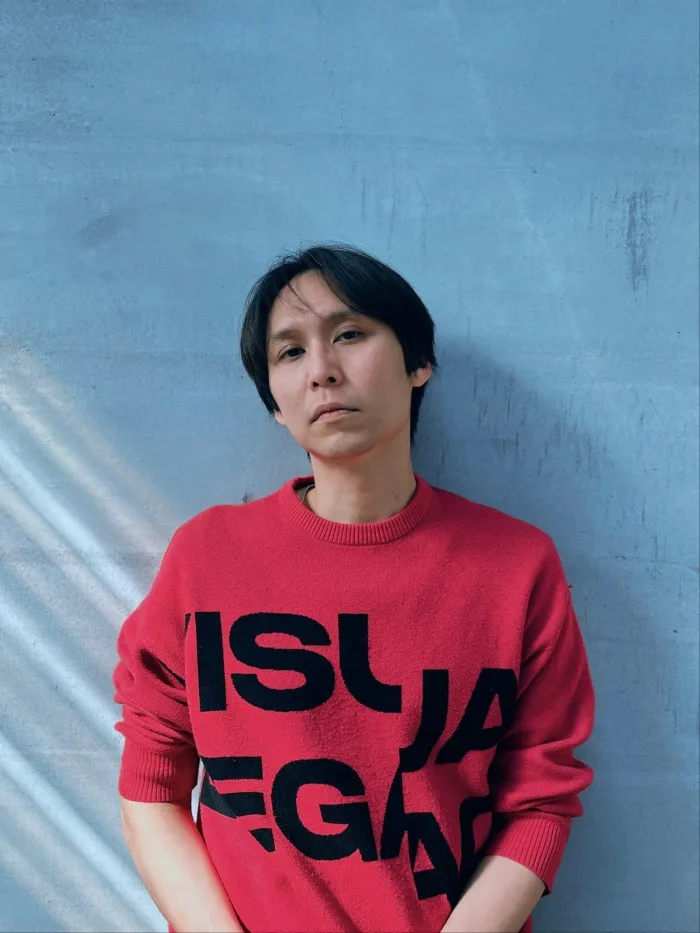 A man with black hair and wearing a fuchsia jumper with black block lettering on the front