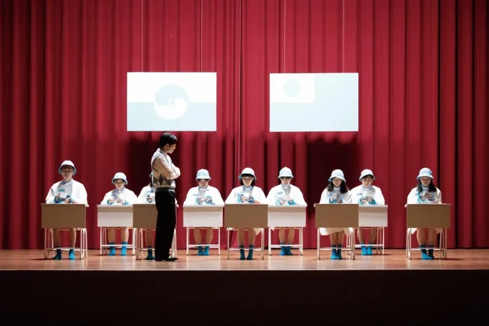 A film still of showing young students at desks arranged on a stage with a red curtain behind them. A man who looks like their teacher stands over them. Two large blue and white posters hang over their heads.