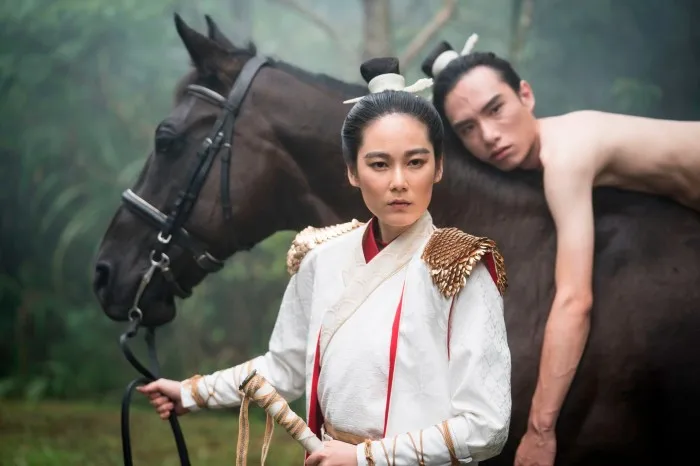 A film still of a woman in a white robe and hair in a bun holds the reins of a horse while a man with the same hairstyle lies on top of the horse.