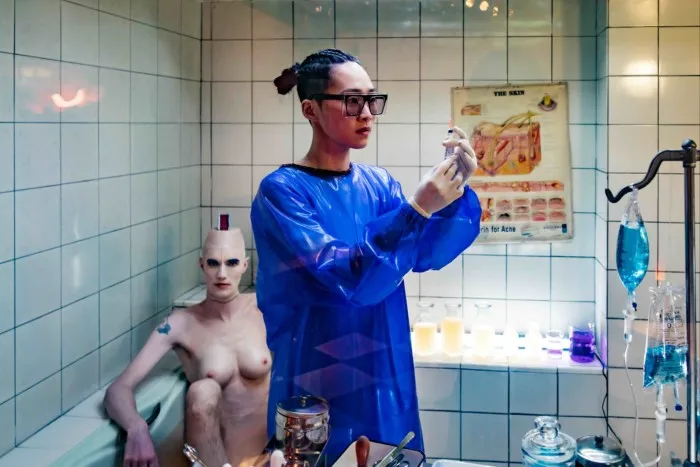 A film still of a woman in blue medical suit inspecting a syringe in a lab. A naked person sits behind her.
