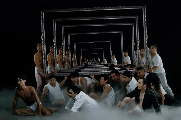A film still showing a group of men crouching on a fog-covered ground, holding a mirror which shows their repeated reflections.