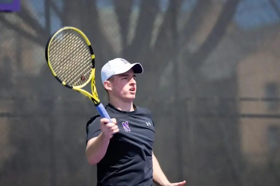 A+tennis+player+in+a+black+shirt+and+a+white+hat+lifts+up+his+racket.