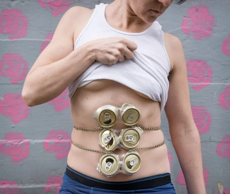 torso with white tank top pulled up to reveal crushed and open six pack of cans where abdominal muscles would be